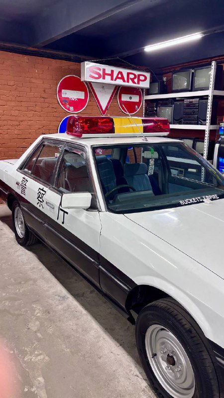 Экспонат внутри музея: белый ретро-автомобиль с мигалкой и рекламным знаком «Sharp» на крыше, размещён в зале с выставочными стендами.