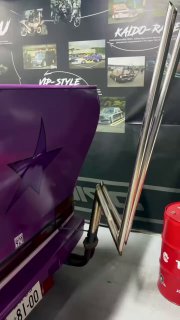 Фрагмент экспозиции: вид на заднюю часть сиреневой машины и рядом расположенные элементы тюнинга, металлические трубы выхлопа и декор зоны музея.
