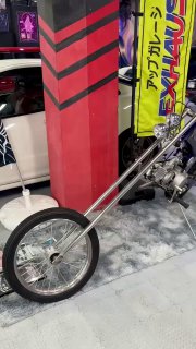 Деталь сцепления и переднего колеса кастомного чоппера или удлинённой передней вилки мото/авто в музее, окружённая яркими стендами и лого Takumi.