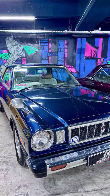 Тёмно-синий классический японский автомобиль с лакированным кузовом, выставочный экземпляр на фоне стенда и других ретро-машин музея.