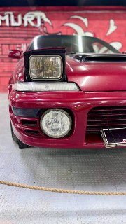 Крупный план передней части красно-бордового автомобиля с иллюстрацией фары и бампера в экспозиции японских ретроавтомобилей в помещении музея.