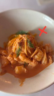 Паста в розовом соусе с кусочками морепродуктов на белой тарелке, фотография помечена крестом, в кадре видна текстура блюда