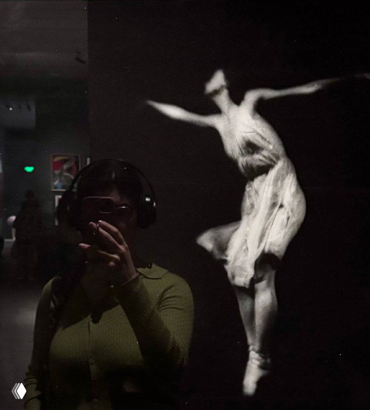 Рефлексия посетителя в витрине на фоне чёрно‑белой проекции танца — человек в наушниках и силуэт танцора на экране позади.