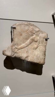 Фрагмент мраморного рельефа I века н.э.: частично сохранившаяся фигура в профиль, установленная на музейной стене с подсветкой.