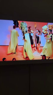 Экран с цветным видео или видеоинсталляцией: сцена с группой людей в костюмах на ярком фоне, часть экспозиции современного танца в музее.