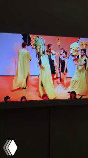 Кадр с экрана: группа артистов на сцене в жёлтых платьях, видеофрагмент перформанса, показанный в зале выставки.