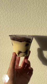 Стаканчик тирамису, снятый сбоку на светлом фоне; видна тёмная шоколадная прослойка и тень руки, минималистичная подача десерта.