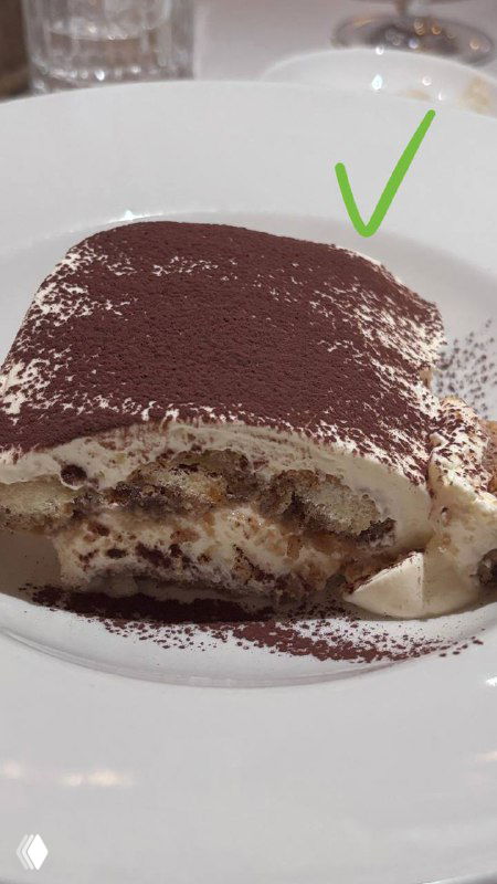 Кусок тирамису на белой тарелке крупным планом, сверху какао-порошок, видно слои крема и пропитанных коржей в разрезе.