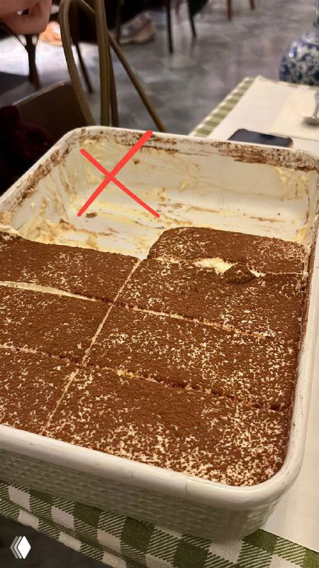 Большой противень тирамису с вырезанными порциями; видно остатки и отметку крестом, иллюстрирующее неудачный исход десерта.