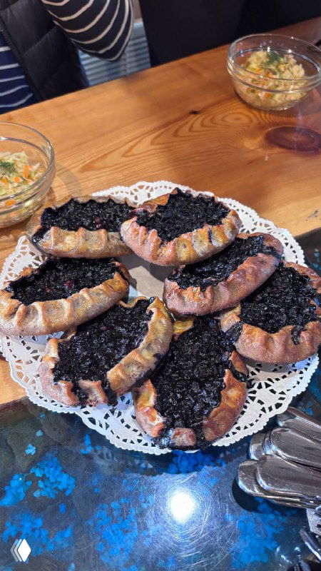 Тарелка с несколькими черничными калитками на кружевной салфетке на деревянном столе; позади миски с финской ухой и столовые приборы.