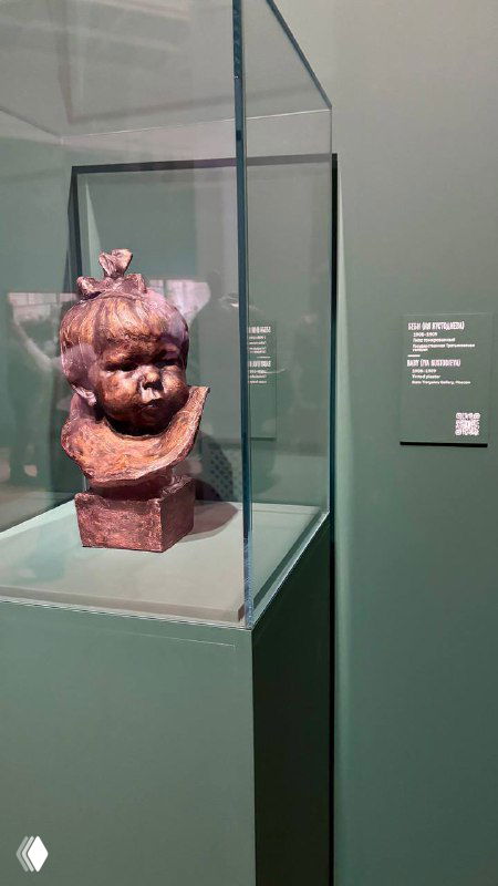 Скульптурный бюст дочери Кустодиева с подписью «бэби», выставленная в музейной витрине рядом с экспонатной табличкой.