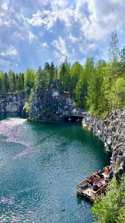 Карьер с лазурной водой между скалами, деревянная платформа с людьми у воды и хвойный лес на фоне при ясном небе.