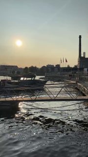 Причал с лодками и промышленными постройками на фоне низкого солнца, отражения на воде и городской силуэт в тёплом вечернем освещении.