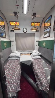 Купе поезда: две полки с подушками и покрывалом, небольшой столик, настенные витражные картины и ретро‑лампы.