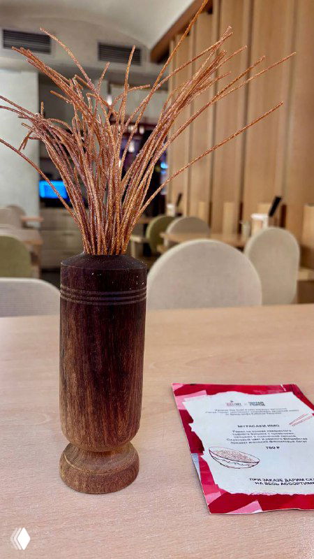 Декоративный деревянный сосуд с высушенными ветками на столе в интерьере Izumi; видны стулья, стол и купон с акцией на фоне.