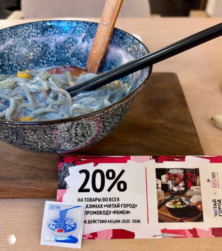 Крупный план фиолетовой лапши, поднятой палочками, рядом виден купон на 20% скидку и деревянная ложка на столе в ресторане Izumi.