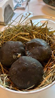 Три чёрных круглых пирожка, поданные в глубокой миске на слое сена и горячих камнях; видна текстура корочки и авторская подача блюда.