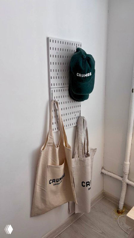 Полотняные сумки и тёмно‑зелёная кепка с логотипом Crumbs, развешенные на перфорированной панели в минималистичном интерьере поп‑ап пекарни