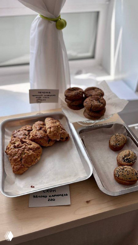 Подносы с ассортиментом печенья, мини‑пирожными и булочками у окна: коричневые печенья и небольшие пирожные на подносах рядом с ценниками