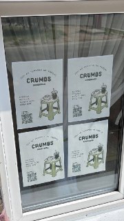 Окно с афишами пекарни: на стекле наклеены несколько листовок с логотипом Crumbs, видны отражения улицы и свет снаружи.