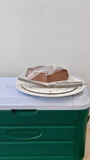 Кусок шоколадного торта Сан‑Себастьян на тарелке, частично закрытый плёнкой, поставленный на пластиковую коробку в простой сервировке.