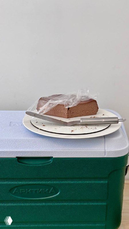 Кусок шоколадного сан‑себастьяна на бумажной тарелке и нож, торт плотной структуры стоит на изотермическом контейнере — демонстрация текстуры десерта