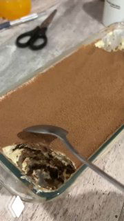 Тирамису в прямоугольной стеклянной форме, поверхность посыпана какао; ложка вынимает порцию, видна текстура крема и слои печенья.