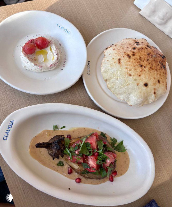 Вид сверху: тарелка с запечённым баклажаном в ореховом соусе, хумус рикотта и надутый горячий лаваш на столе в ресторане Claudia, логотип на посуде.