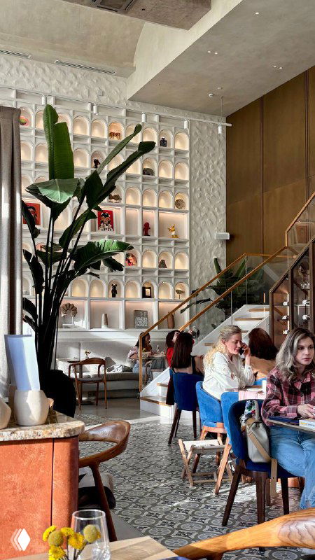 Интерьер ресторана Claudia с высоким потолком, декоративными стеллажами и растениями, гости за столиками и мягкие кресла синего цвета.