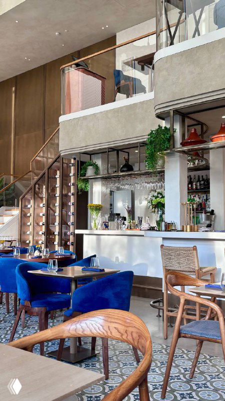 Барная зона и лестница в ресторане Claudia: открытая планировка, синие кресла, столы и элементы декора на фоне кухонной зоны.