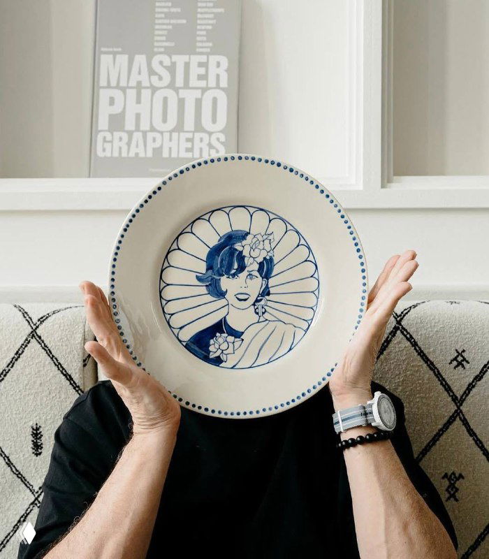 Рука держит декоративную тарелку с иллюстрацией на фоне дивана и интерьерных постеров — акцент на посуде и дизайне ресторана Claudia.