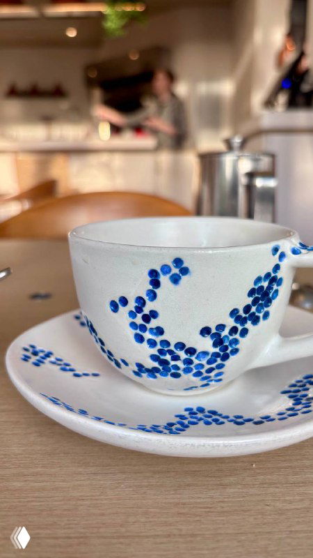 Крупный план керамической чашки с синим узором на блюдце, на заднем плане стол и интерьер ресторана Claudia — акцент на посуде бренда.