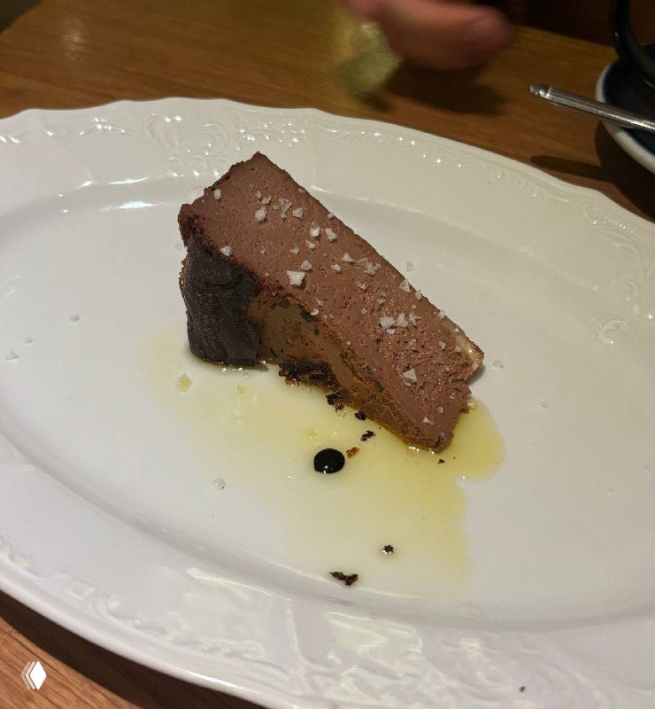 Кусок шоколадного баскского чизкейка на белой тарелке, посыпанный морской крупной солью, с каплями оливкового масла и бальзамиком.