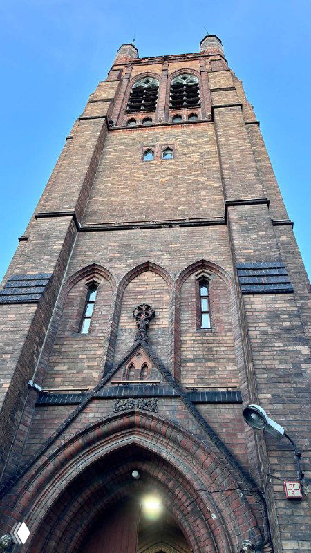 Высокая кирпичная колокольня англиканской церкви Св. Андрея в Москве, вид снизу вверх с готическими арочными окнами и каменной отделкой фасада.