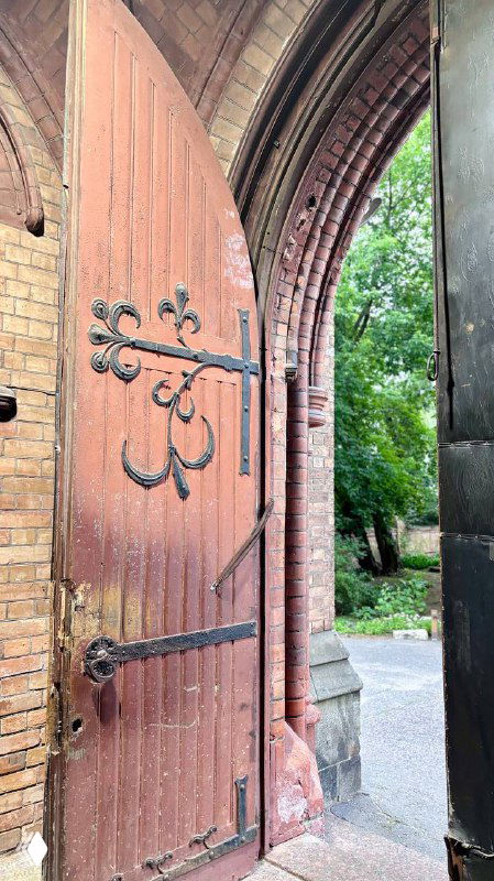 Открытая часть деревянной двери церковного портала с видом на зелёную аллею позади здания и часть кирпичной кладки у входа.