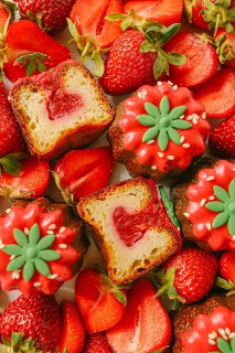 Несколько канеле с красной клубничной начинкой и зелёными декоративными элементами, расположенные на подложке из свежих разрезанных клубник.