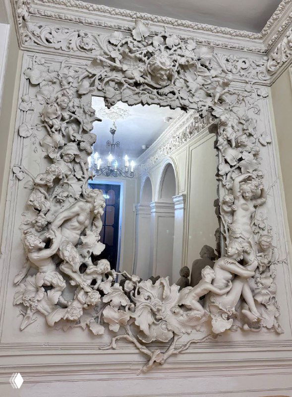 Крупный план: массивная резная белая рама зеркала с обильной лепниной, ангелами и растительными орнаментами в интерьере усадьбы.