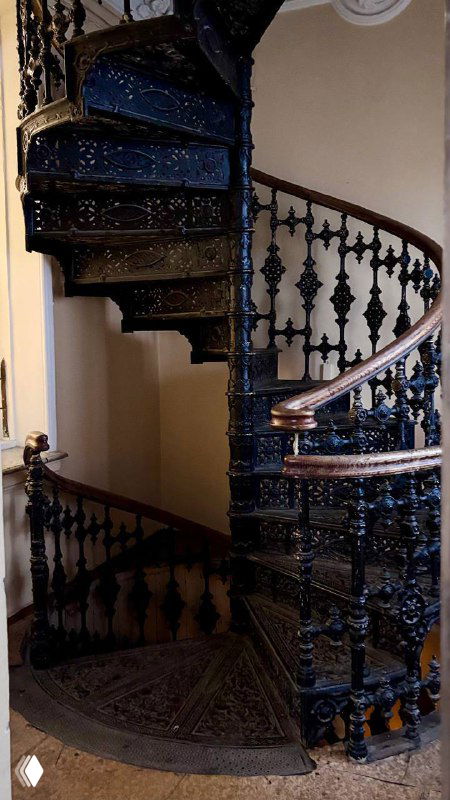 Чёрная винтовая чугунная лестница с декоративными балясинами, ажурными элементами и тёмным деревянным поручнем в историческом интерьере усадьбы.