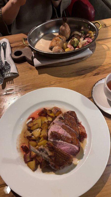 Крупный план тарелки с утиным филе‑супрем, кусочки яблок и картофель, соус на белой тарелке на деревянном столе рядом с чашкой и прибором.