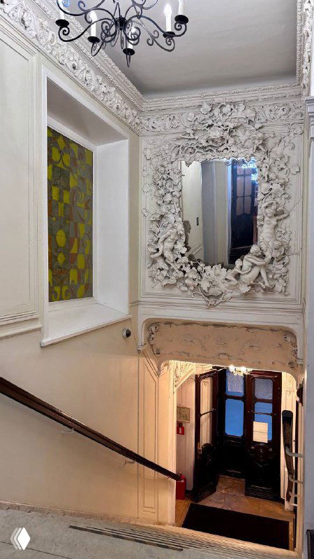 Ракурс лестничного холла усадьбы: большое резное зеркало, витражное окно, классическая люстра и декоративные орнаменты в одном кадре.