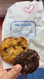 Две печеньки, тёмная и светлая, в руке на бумажном пакете с логотипом METEORITE, видна текстура, крошки и уличный фон.