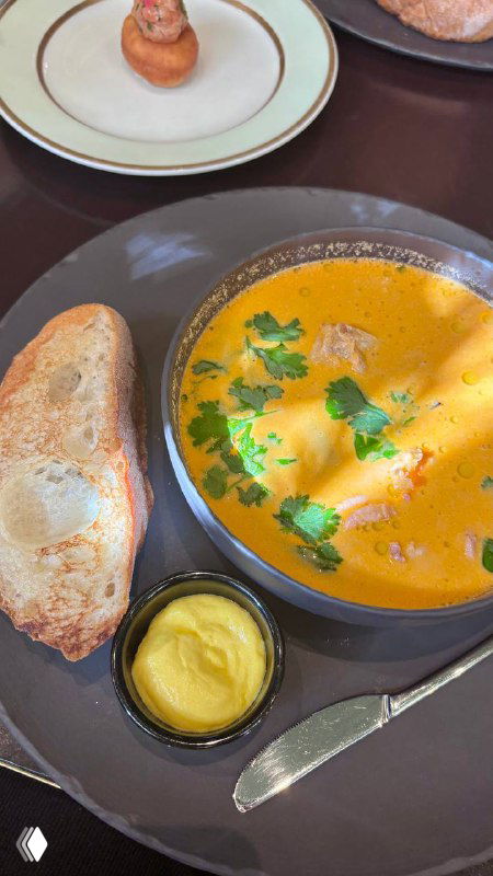 Тарелка с ухой в Geraldine: кремовый оранжевый суп, зелень, кусочки хлеба и небольшая ёмкость с соусом — подача блюда на темной тарелке.
