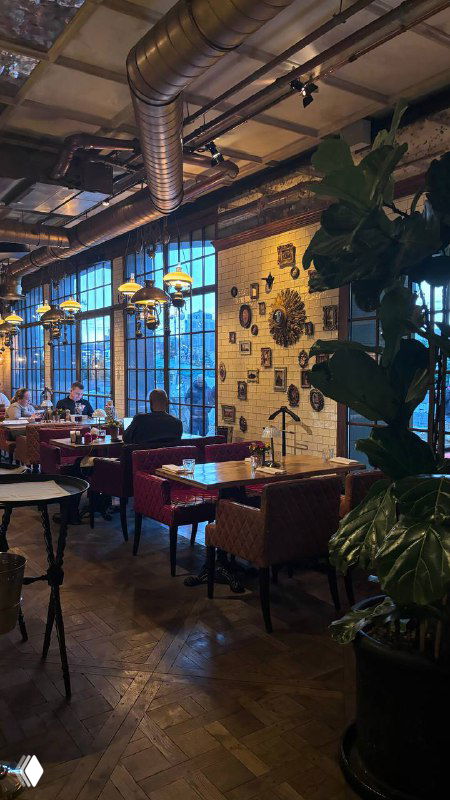 Обеденная зона у больших окон с плиточной стеной и лампами, столы и стулья в вечернем освещении, растительность в кадре.