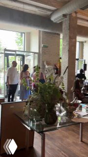 Другой ракурс зала бистро: у входа стоят люди, стеклянные столы и цветочные композиции на фоне кирпичных стен.