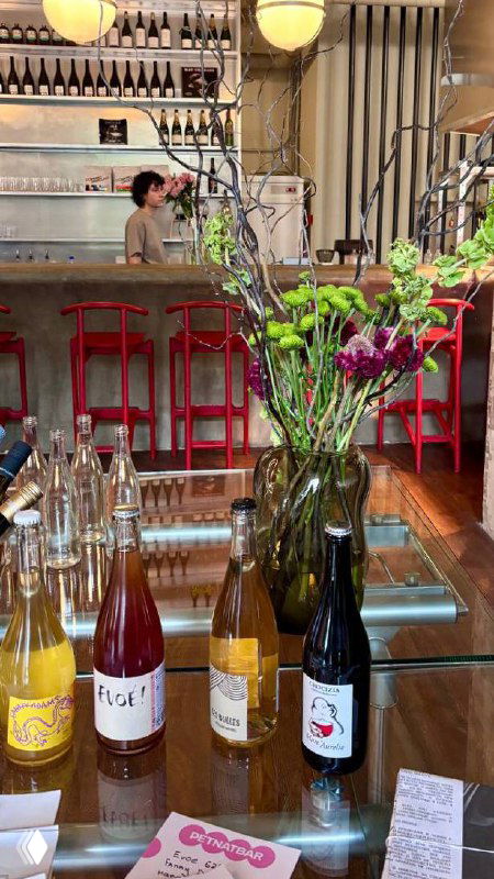 Барная стойка и витрина с бутылками лимонадов: стеклянные бутылки, букеты цветов и часть красного барного стула на переднем плане.