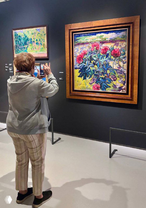 Посетитель фотографирует картину со смартфона в галерее: женщина стоит перед яркой цветочной работой, видно пол и стенды музея.
