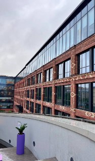 Вид наружного фасада здания музея с кирпичным оформлением и большими окнами, снятый с террасы; в кадре часть балкона и городской фон за музеем.