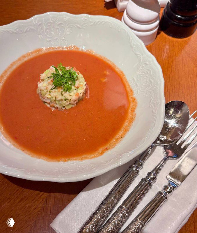 Тарелка гаспачо с центральным шариком гарнира, рядом сложенные серебряные столовые приборы и салфетка на деревянном столе ресторана.