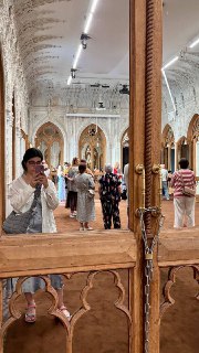 Зал с зеркалами, посетители отражаются в мебели и витринах; видно множество людей на экскурсии и резные архитектурные детали.