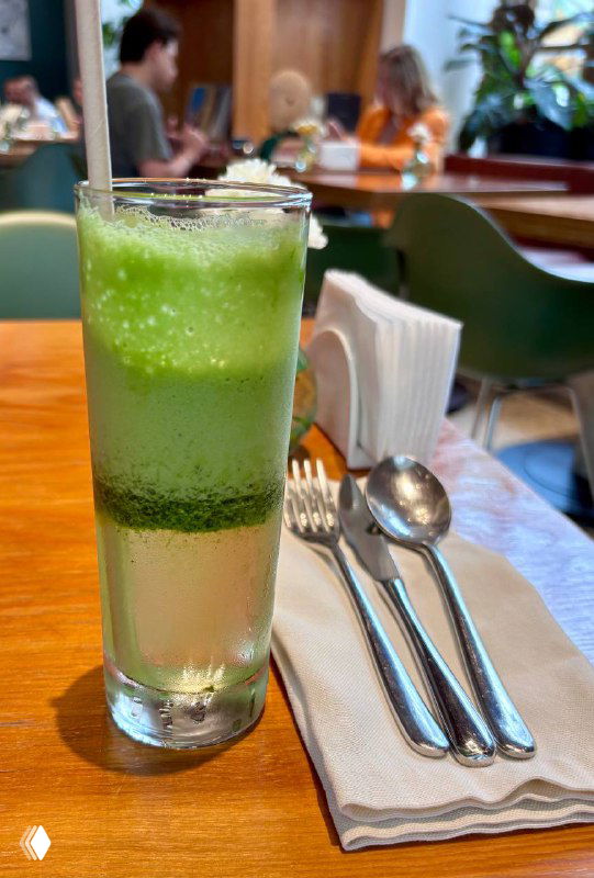 Высокий прозрачный бокал лимонада щавель-алоэ с заметным большим куском льда, на фоне приборов и интерьера кафе Flaner.